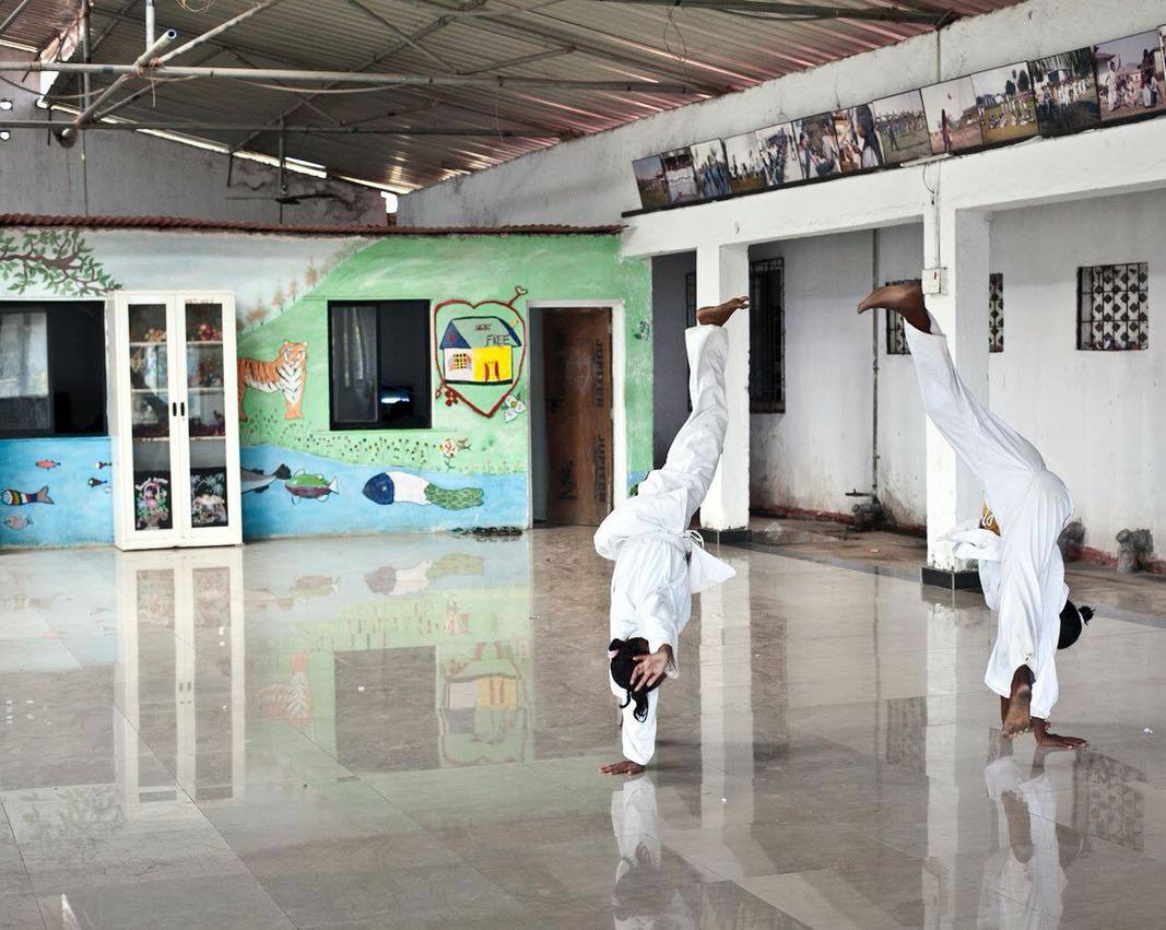 Two girls turning handstands in the bright hall of a Rescue Foundation shelter home, beside a wall mural of a tiger, fish and a house.